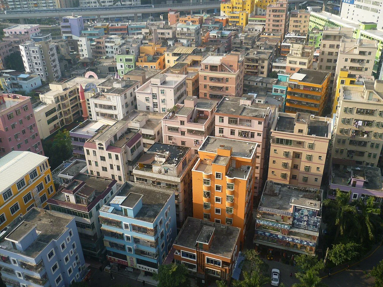 Aerial daytime view of Dafen Oil Painting Village showing the dense cluster of low-rise buildings painted in a rainbow of colors — yellow, blue, pink, orange — that characterizes the village's distinct visual identity