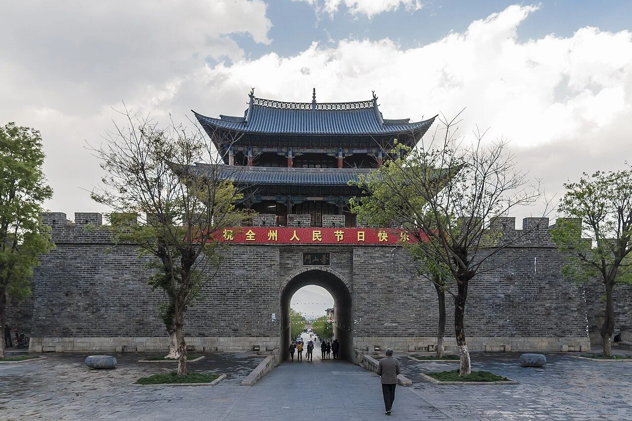 The West Gate tower of Dali Old Town — the iconic 600-year-old gate tower of the walled city, with grey-brick city wall, people walking through, and a red national-day banner above the gate arch
