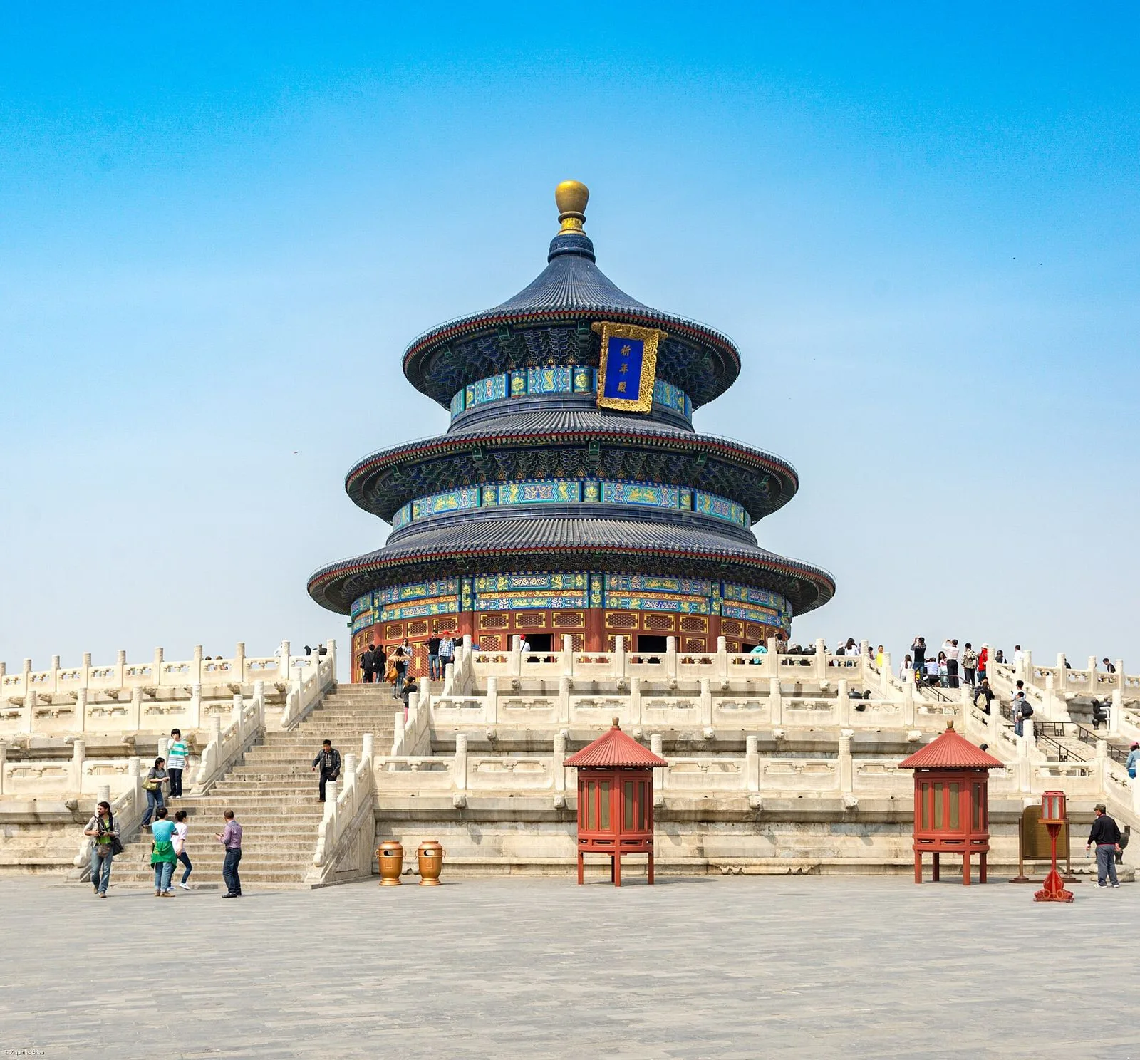 Wide view of the Hall of Prayer for Good Harvests from the steps, with dozens of visitors visible on the platform and stairways, the full white-marble base and blue-roofed superstructure of the building rising above, and red lanterns on tall stands in the foreground — the scale of the Hall of Prayer as a working public space, with tour groups and individual visitors sharing the same experience