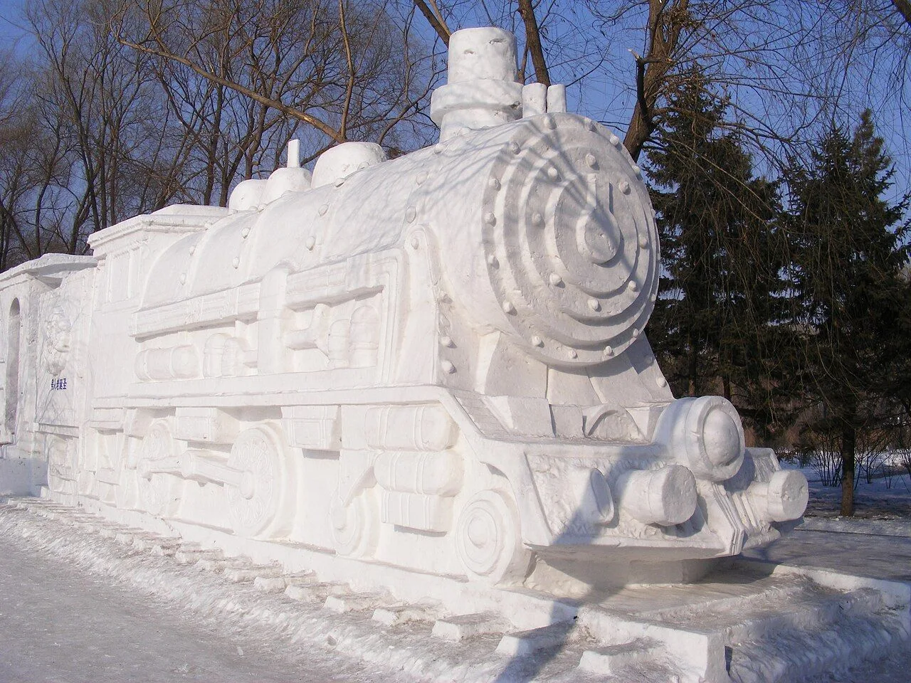 A massive snow sculpture of a steam locomotive at the Harbin International Ice and Snow Sculpture Festival, daytime view with pine trees — the 100% snow (not ice) sculptures are built by teams from around the world and can be 20+ meters tall