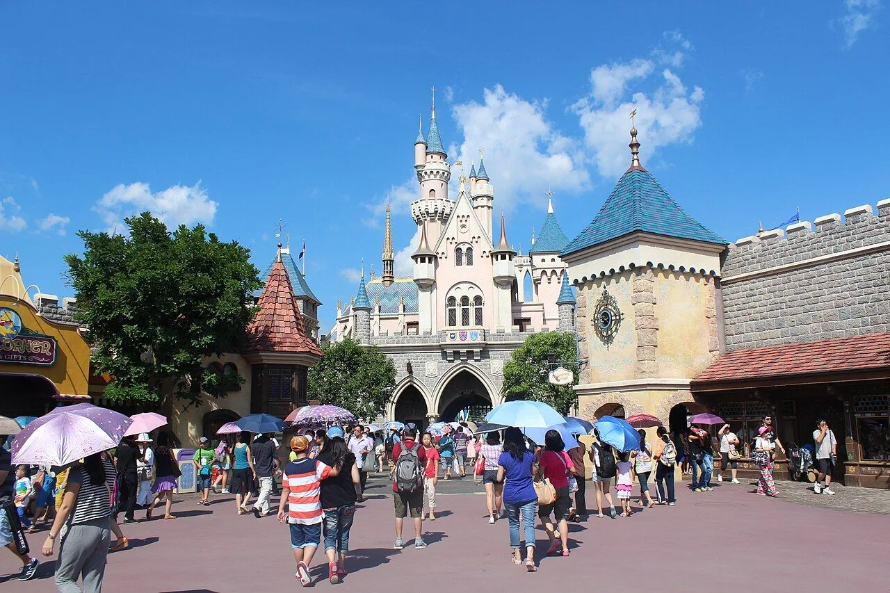 Main Street at Hong Kong Disneyland with visitors walking toward the Sleeping Beauty Castle under a sunny sky — the iconic approach to the park, with the colorful shop fronts and the castle rising in the background