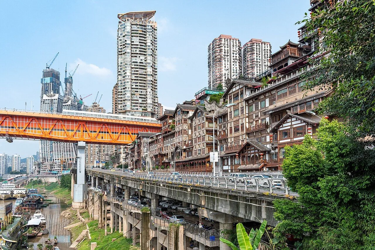 Daytime view of the Hongya Cave stilted-house complex along the Yangtze tributary in Chongqing — the 11 traditional wooden tiered buildings rising from the riverbank, with a road bridge and the modern high-rises above, showing the 600-year history meeting the 21st century