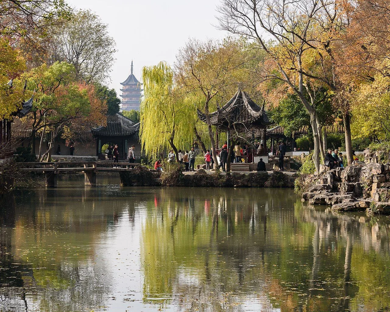 The central pond of the Humble Administrator's Garden (拙政园) in Suzhou, with willow trees and a traditional pavilion reflected in the water; the North Temple Pagoda (北寺塔) of Suzhou is visible in the background — the typical "borrowed scenery" (借景) technique in Chinese garden design