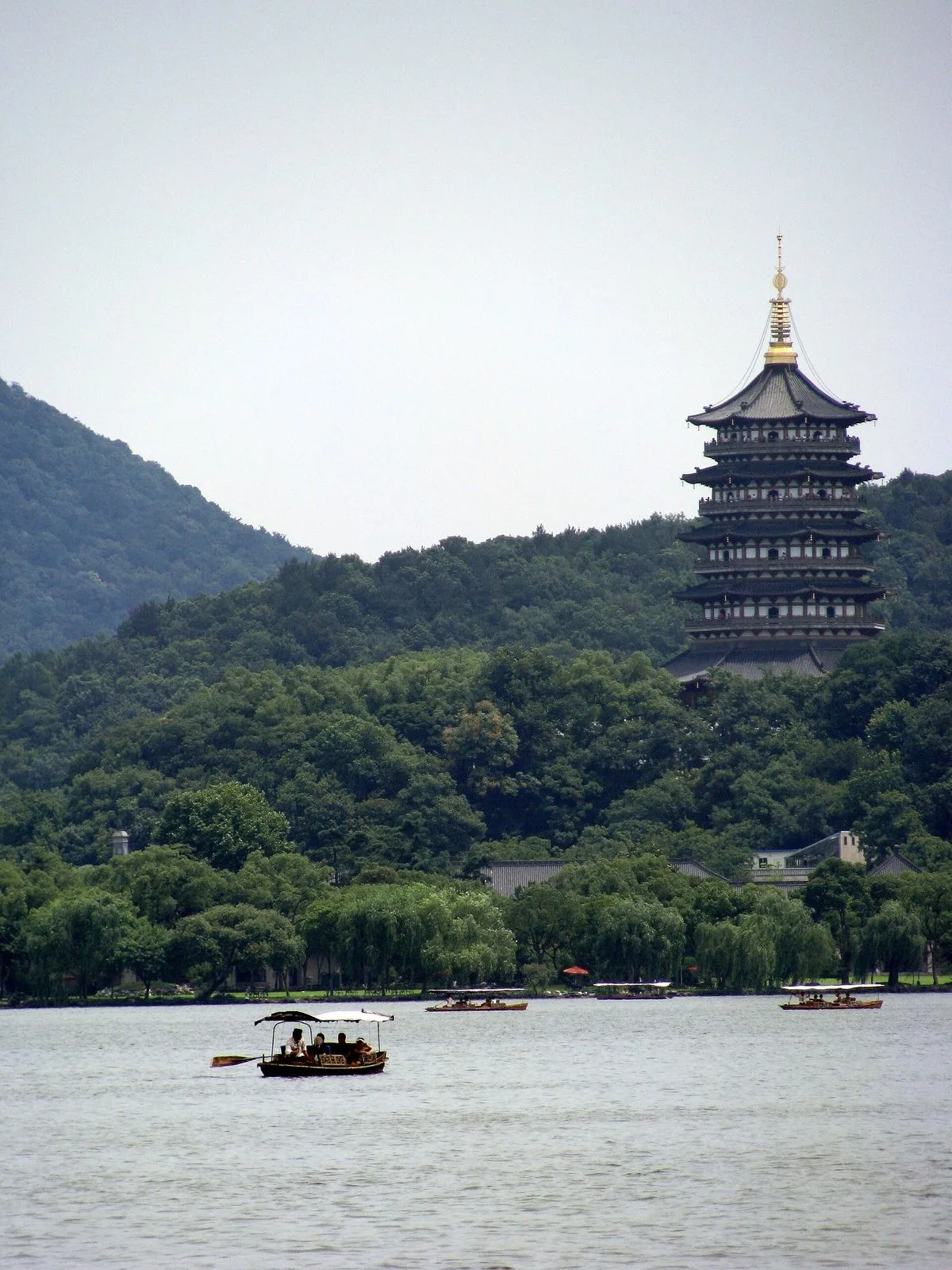 Wide view of West Lake with the Leifeng Pagoda on the southern shore in the distance, a traditional wooden boat with canopy and rower in the foreground, and a smaller boat further out on the calm water — the classic Hangzhou scene of pagoda-by-the-lake with the iconic hand-rowed boats that have carried visitors across West Lake for over a thousand years