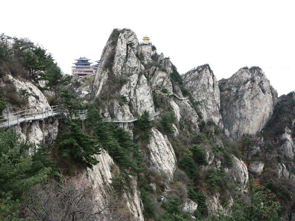 The wide mountain landscape of Laojun Mountain in Henan — two pavilion complexes sitting on the granite peaks, a winding plank road clinging to the cliffs, pine forest filling the valleys, and the dramatic topography that gives the mountain its status as a sacred Taoist site (named after Laozi, the founder of Taoism)
