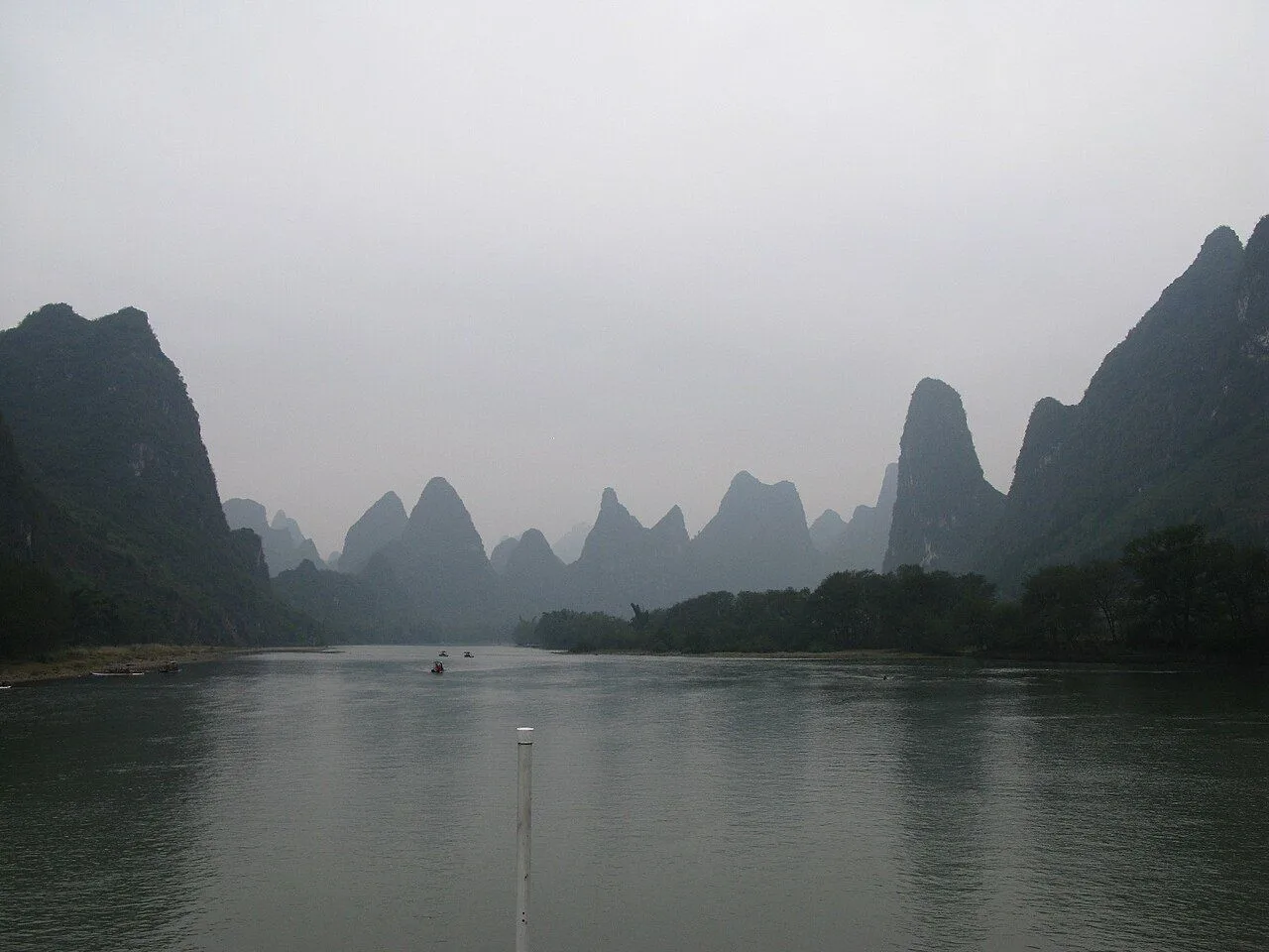 A wider view of the Li River cruise from Guilin to Yangshuo — multiple tour boats on the calm water, layers of karst peaks fading into the misty distance, and a single bamboo pole in the foreground; the classic 84-km journey through 5 counties of Guangxi province
