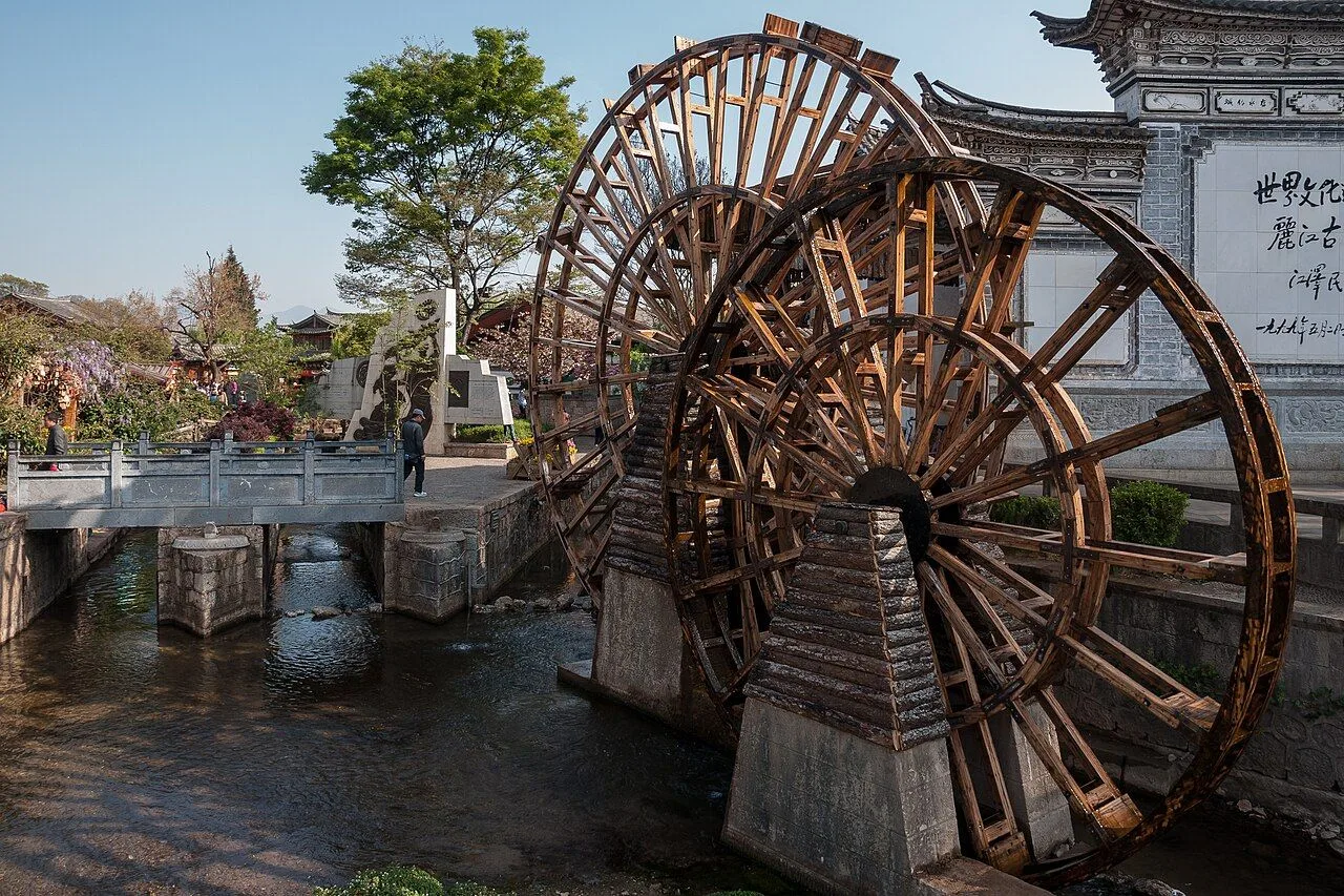 The iconic water wheels on the canal at the entrance to Lijiang Old Town in Yunnan — with the stone bridge and traditional Naxi architecture in the background, the symbol of the town and its 800-year-old water-powered economy fed by the Jade Dragon Snow Mountain snowmelt