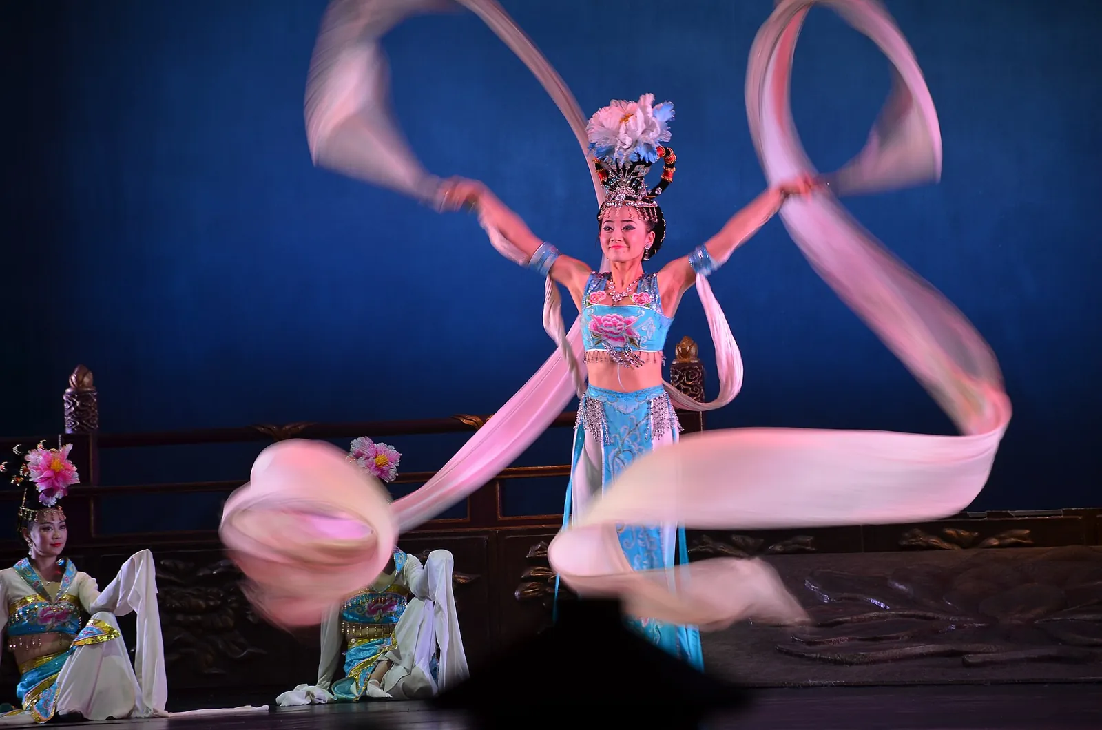 A Tang Dynasty show dancer in pink and blue silk with trailing sleeves, mid-motion on stage at Tang Paradise, with two seated performers in Tang-style white and gold silk in the background
