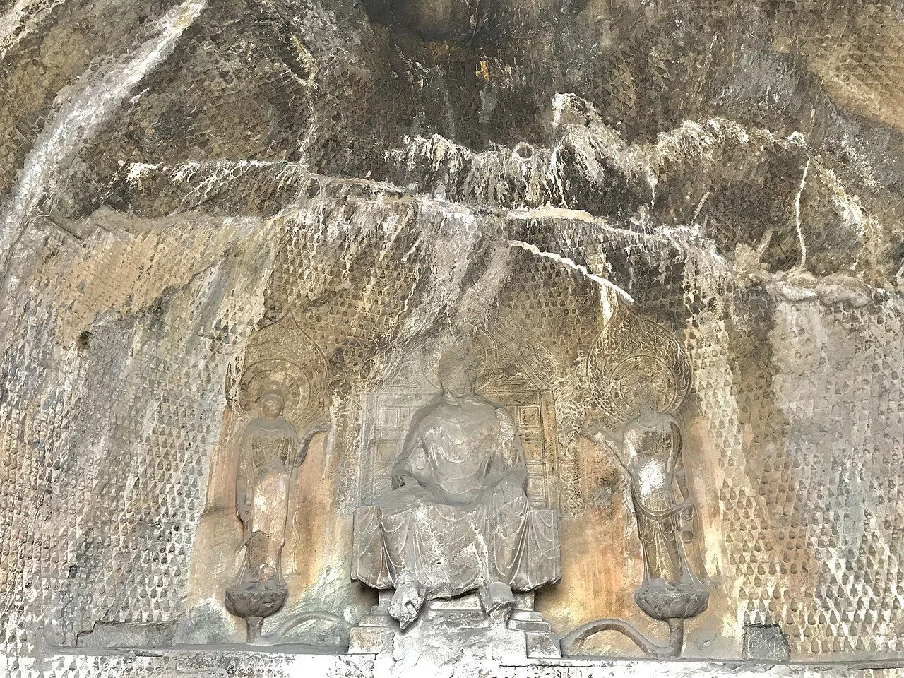 The interior of a Longmen cave niche showing a Tang dynasty Buddha triad — a central seated Buddha flanked by two standing bodhisattvas, surrounded by hundreds of smaller carved figures covering the wall; the pinnacle of Tang dynasty Buddhist sculpture from the 7th-8th century