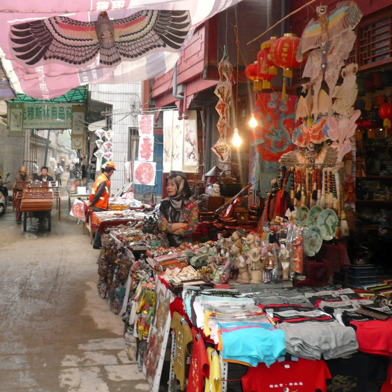 A street vendor in the Xi'an Muslim Quarter — Hui Muslim woman surrounded by colorful kites, embroidered textile souvenirs, paper cutouts, and traditional Chinese lanterns, the diverse crafts sold along the bazaar street that has traded since the Ming dynasty. Most vendors are Hui Muslim with their own craft traditions