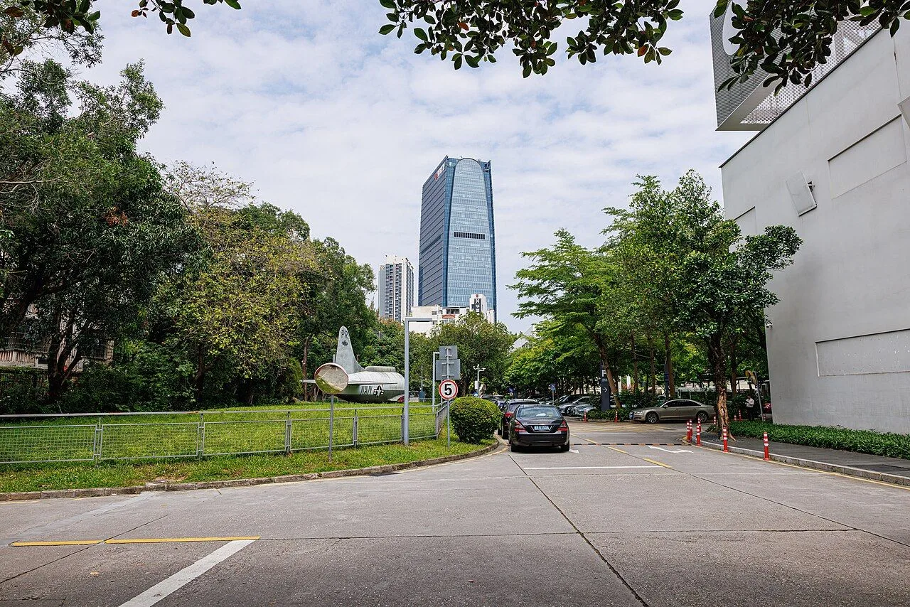 A vintage propeller plane displayed as a public art piece at OCT LOFT in Shenzhen, with the modern Nanshan skyscraper skyline visible in the background — the contrast of industrial heritage and the new Shenzhen tech district. The complex was originally a 1990s electronics manufacturing plant