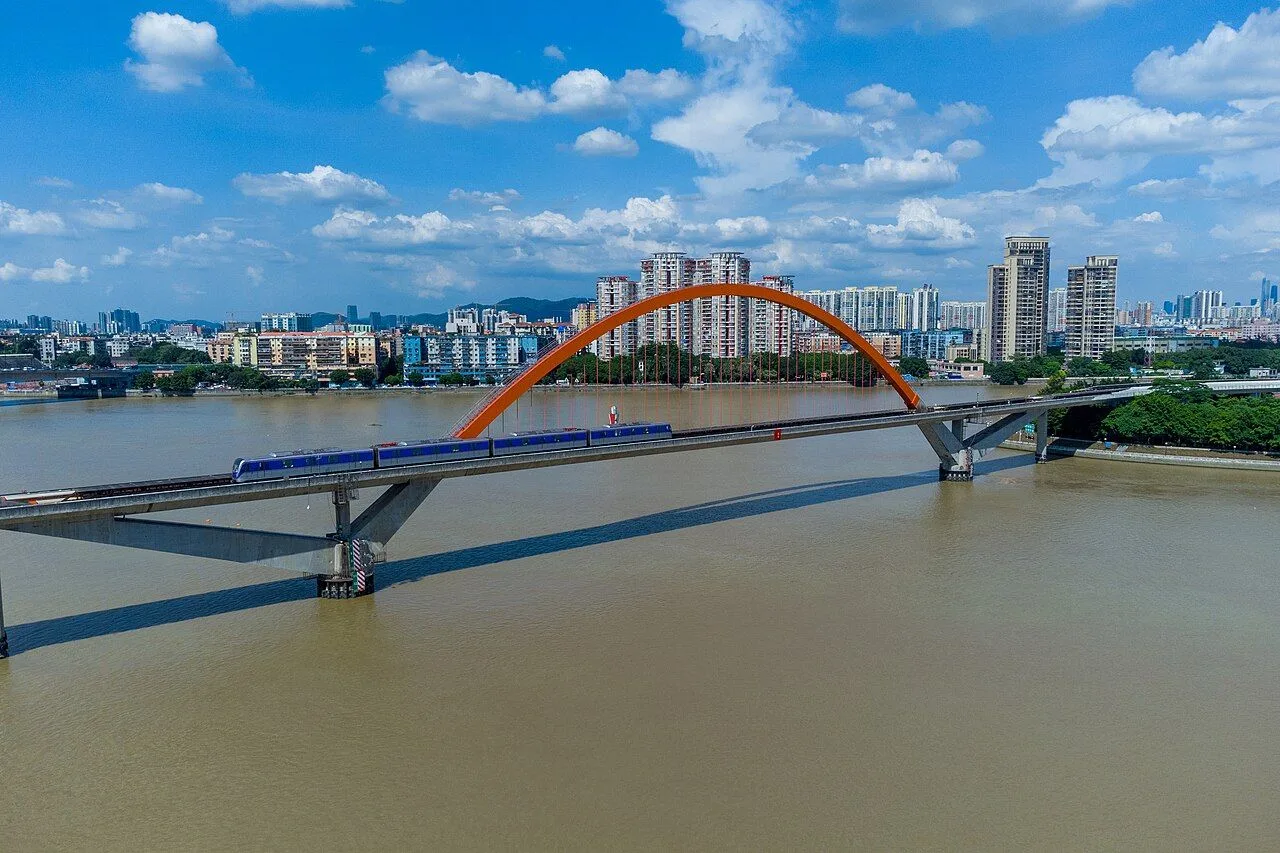 A wider daytime view of the Jinshazhou Pearl River Bridge with its signature orange arch and a metro train crossing, showing the full bridge profile over the wide brown river with the dense Liwan residential towers and distant mountains visible in the background