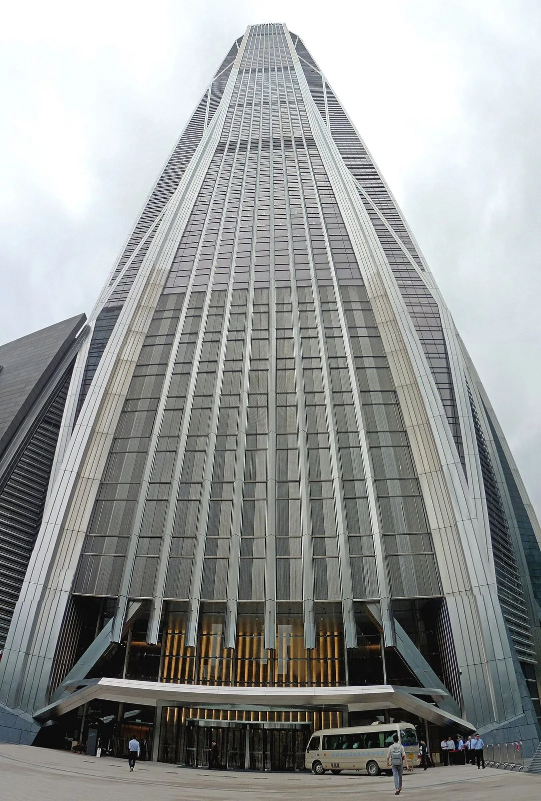 A dramatic low-angle view looking straight up at the base of the Ping An Finance Centre, the tapered glass facade rising vertically to the tapered crown, with the building's full 599m height compressed in the frame and a small white minibus parked at the entrance giving a sense of scale to the monumental structure