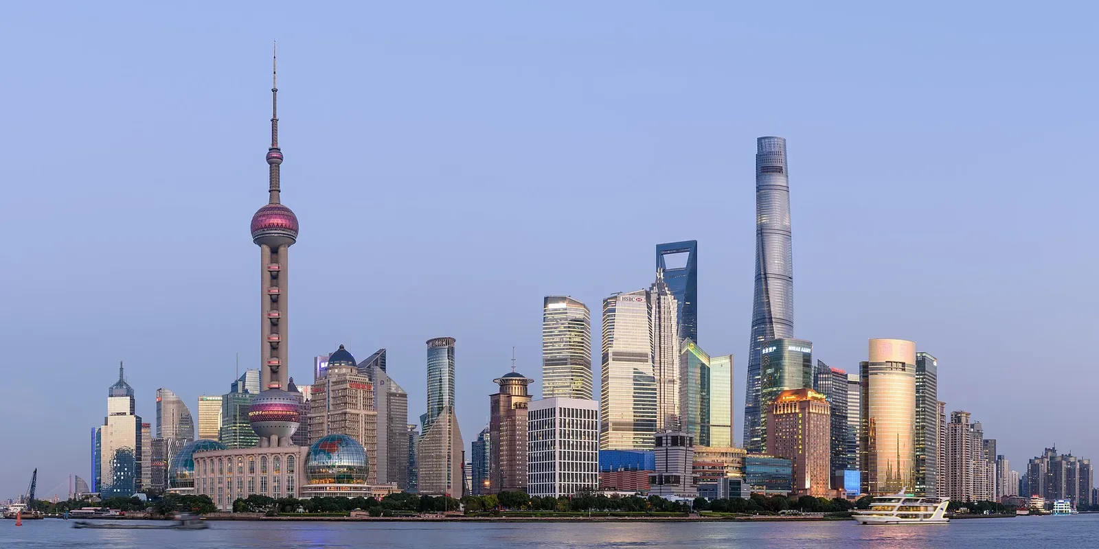 The Pudong skyline seen from the Bund during daytime — Oriental Pearl TV Tower, the Shanghai World Financial Center, Shanghai Tower, and Jin Mao Tower lined up along the river, with the Huangpu River in the foreground and a pale blue sky above