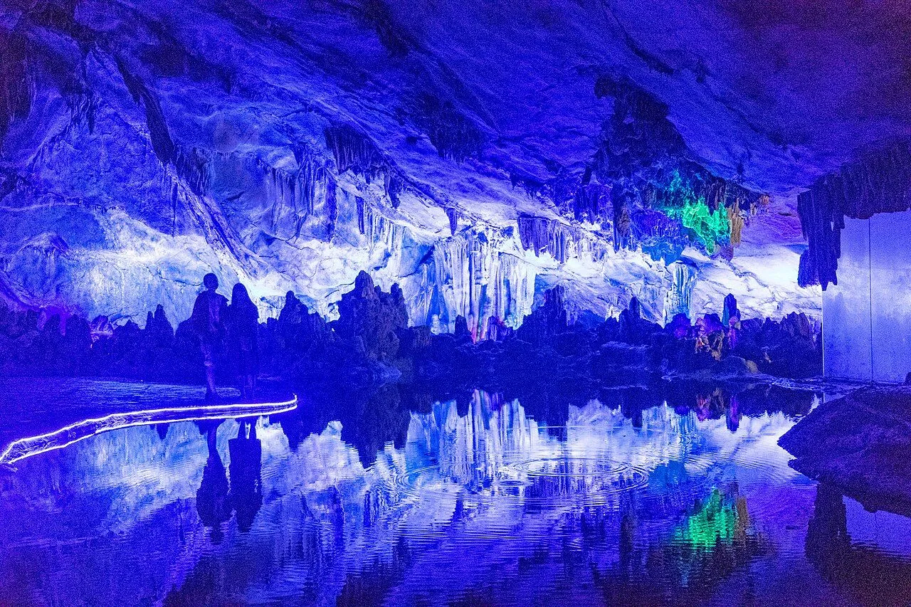 A blue-lit chamber inside Reed Flute Cave — visitors walk along a raised pathway above a still underground pool that mirrors the dramatic stalactites hanging from the ceiling, the cave’s most photogenic spot and one of the most-captured cave interiors in China, where a single drop of water on the surface creates a single perfect ripple in the reflection