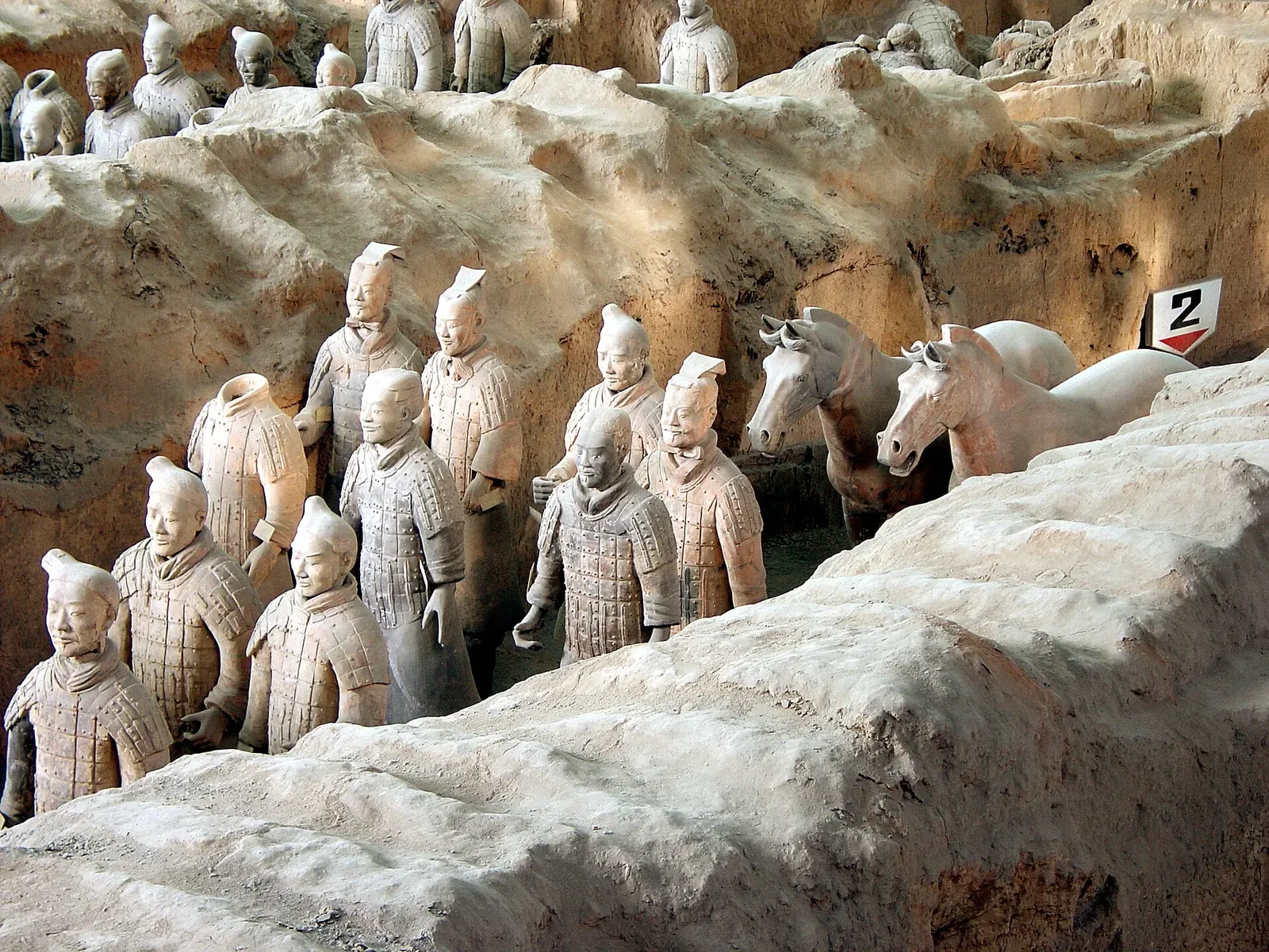 Rows of terracotta warriors standing in formation inside Pit 1 at the Terracotta Army museum, with the earthen trench walls visible behind them and a few horse figures on the right