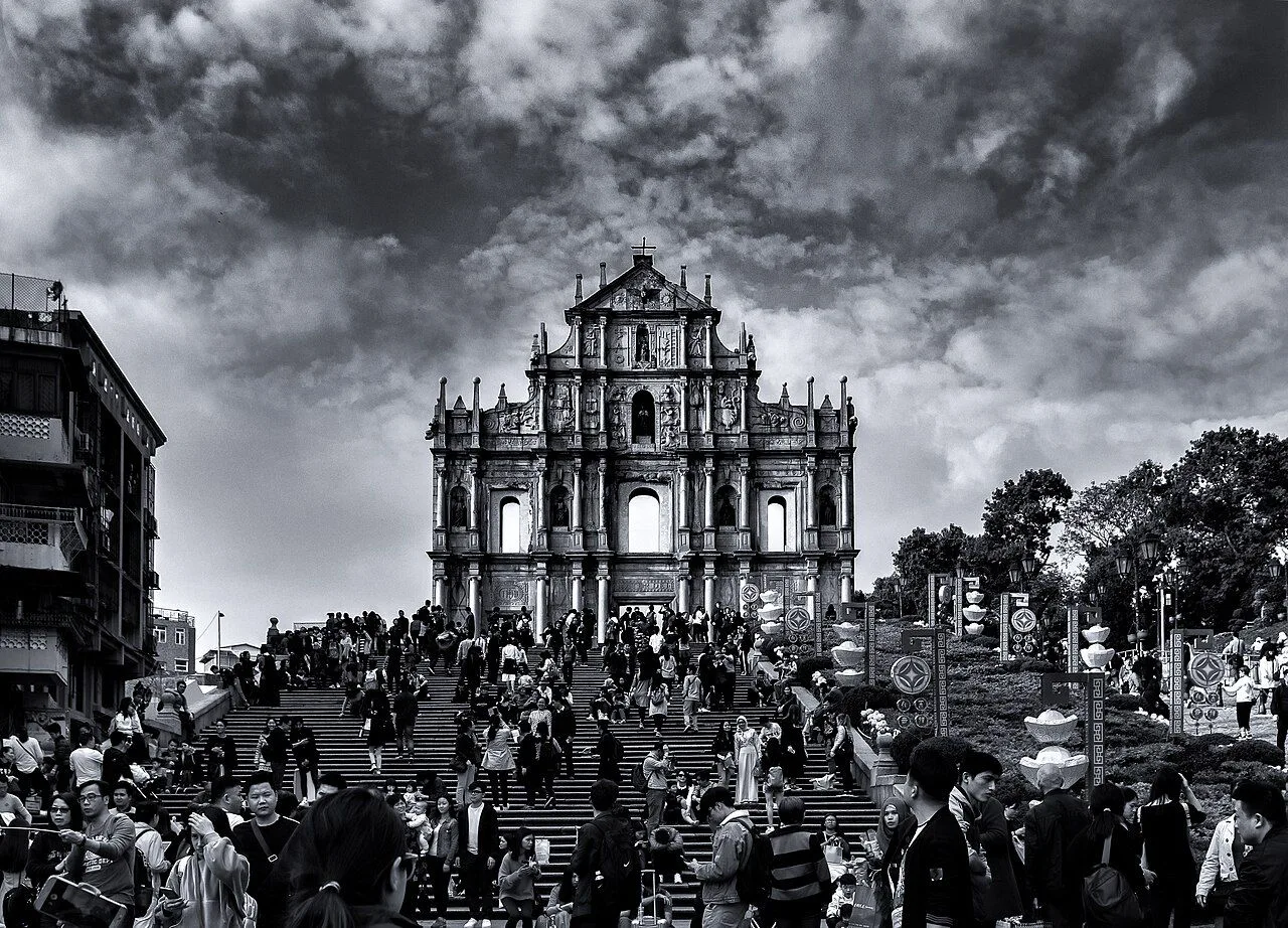 The Ruins of St Paul’s facade in dramatic black-and-white — the carved Baroque stone front rises above a long flight of stone steps crowded with visitors, the entire Macau skyline visible behind, the image that appears on the Macau 5-pataca banknote and is the universal symbol of the city, where the great fire of 1835 left only the granite facade standing