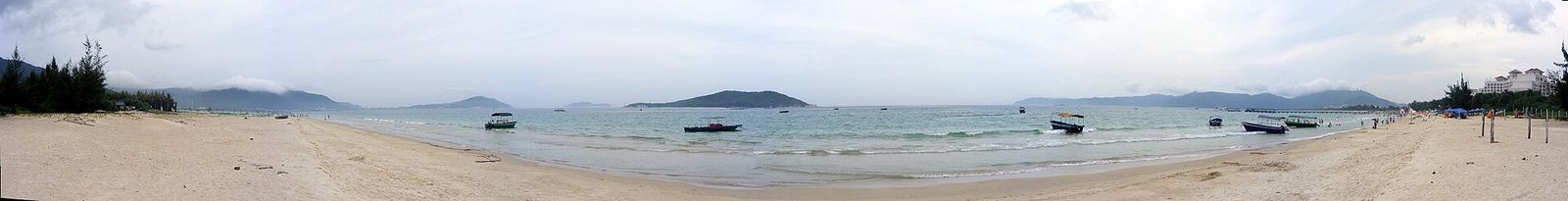 A panoramic view of Yalong Bay (亚龙湾) — the crescent-shaped bay with anchored fishing boats, white sand beach, the forested headlands of the tropical coast, and luxury resort buildings visible at the far right, the bay that was selected as one of the world’s top 10 best beaches by China National Tourism in 2001