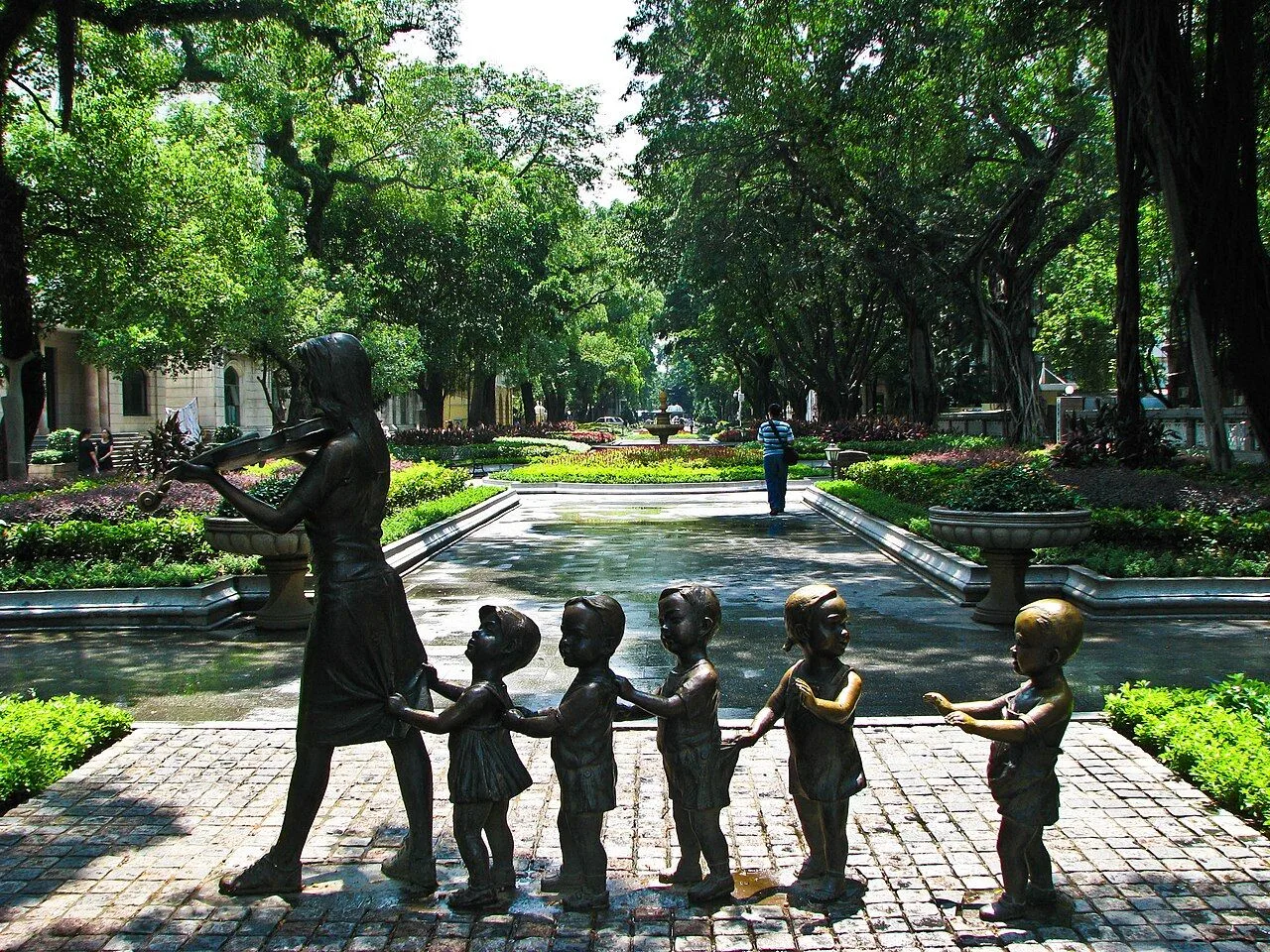 A bronze sculpture of a teacher leading a line of children on a quiet pedestrian path inside Shamian Island, set among lush tropical greenery and the manicured paths of the island's central park, with a Chinese visitor walking in the background