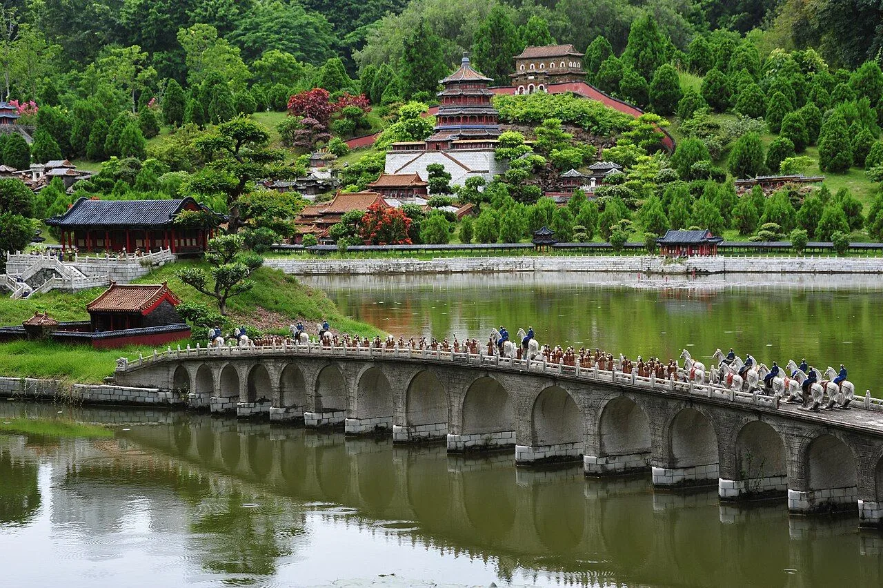A 1:15 scale miniature replica of the 17-arch bridge at the Summer Palace in Splendid China, with rows of tiny Chinese figures in traditional dress crossing the multi-arched stone bridge over the model lake, miniature pavilions and imperial buildings on the far bank under bright midday sun