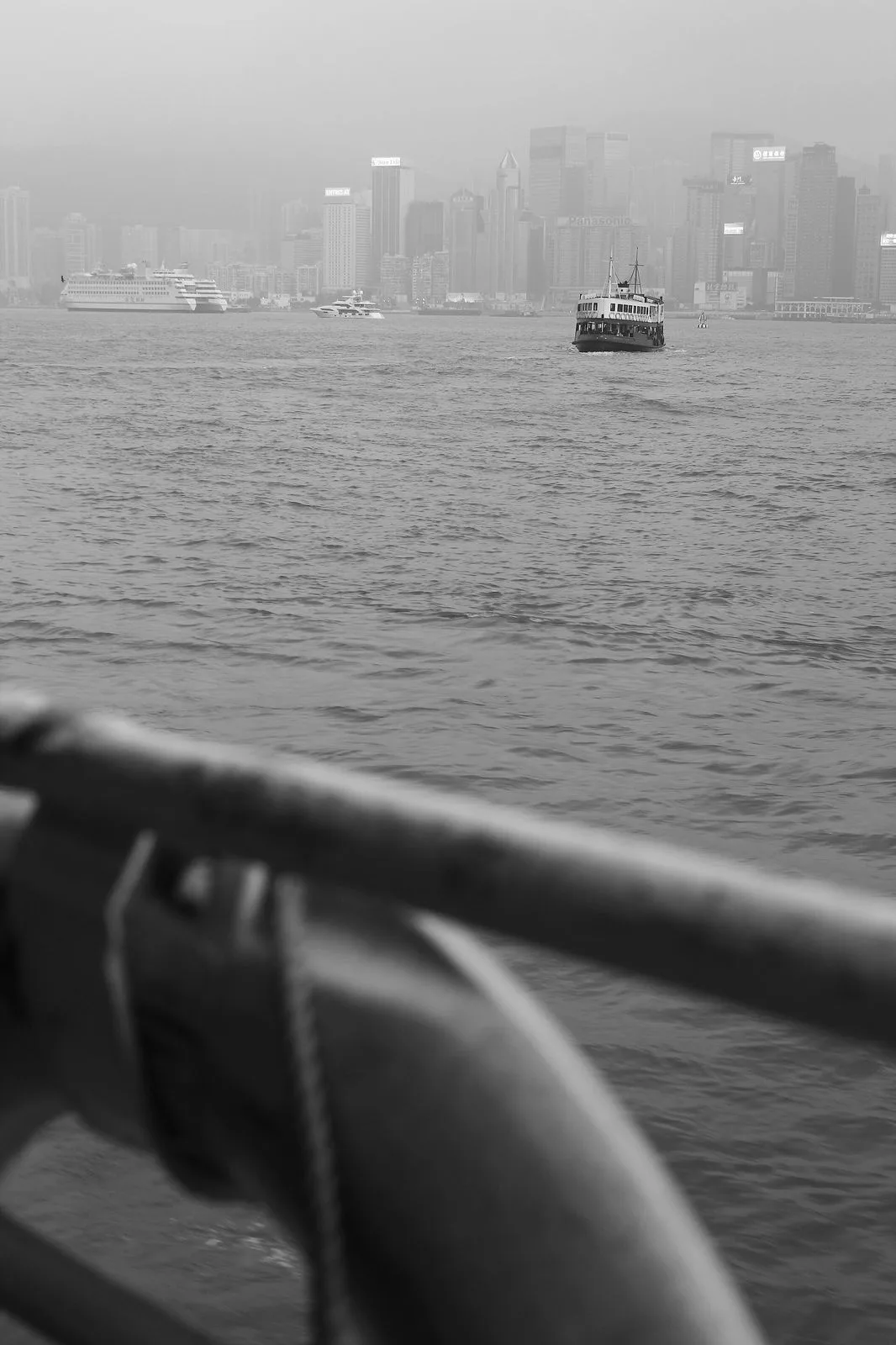 A view from the upper deck of a Star Ferry crossing Victoria Harbour — a dramatic black-and-white shot of the Hong Kong Island skyline in the misty distance, the ferry’s own railing curving into the foreground, the perspective the 4.2 million annual Star Ferry passengers experience on the 7-minute crossing between Kowloon and Hong Kong Island
