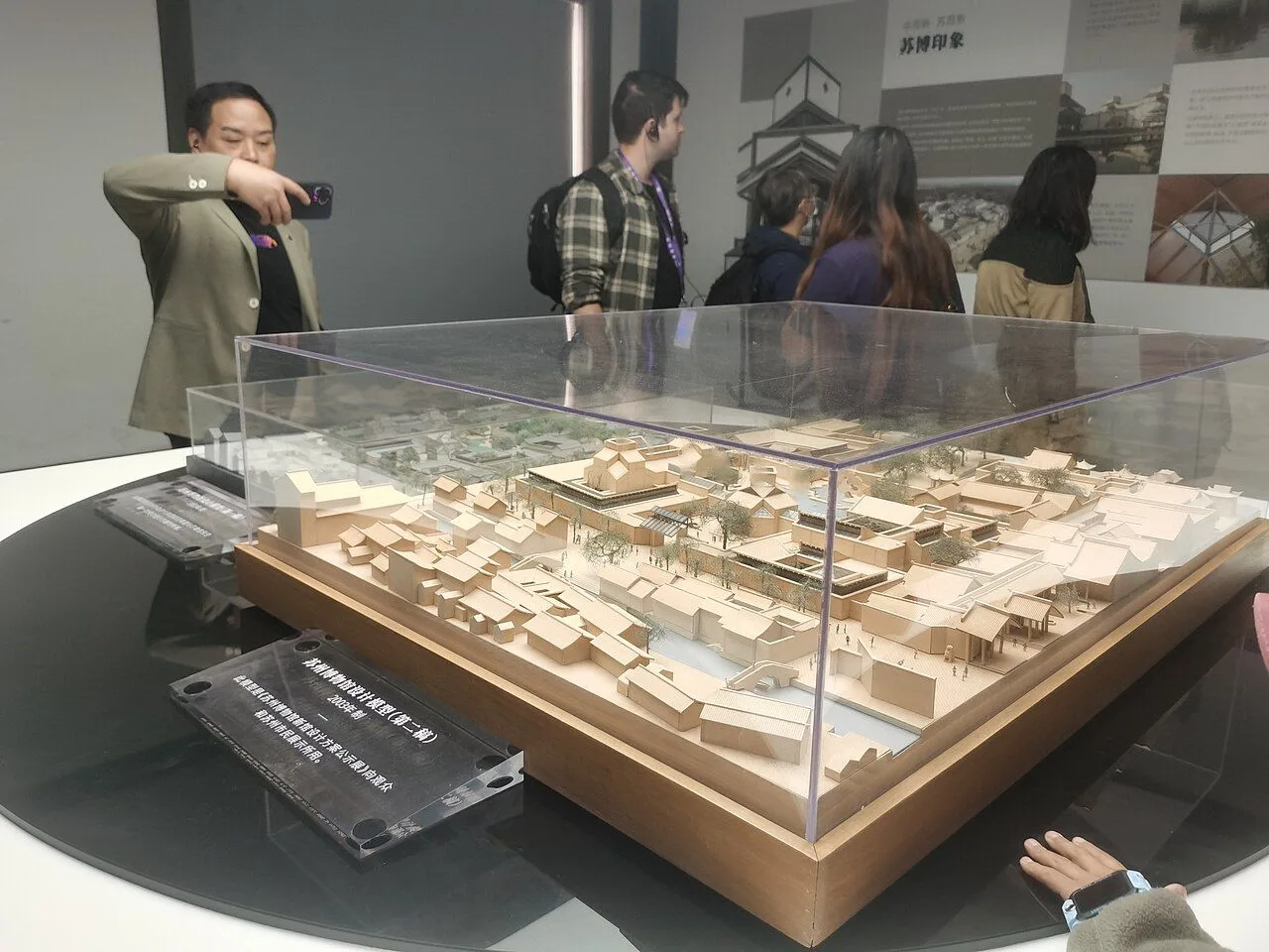 The original architectural design model of the Suzhou Museum by I.M. Pei on display inside the museum, with a Chinese visitor in a green jacket photographing the model with a smartphone, showing the precision miniature of the building and grounds