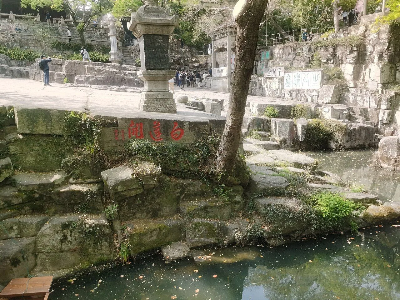 The Bailiankai (Thousand Lotus Petals) carved stone spring at Tiger Hill, Suzhou, with a stone stele in the center and traditional Chinese characters carved in red on the rock face, with a small pool of water below and visitors climbing stone steps in the background