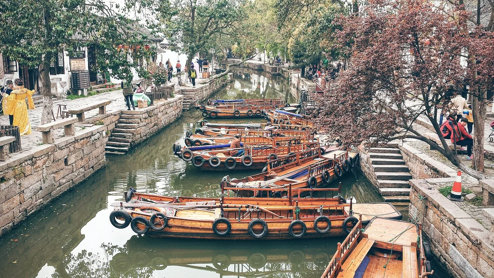A row of traditional wooden boats moored along the Tongli canal — the black-tiled roofs and white-washed walls of Ming-and-Qing-era merchant houses lining both sides of the waterway, the boats with curved canopies used to take visitors on the 30-minute canal cruise through the old town, the everyday scene of daily life that has continued here for 600 years since Tongli was founded in the Song dynasty