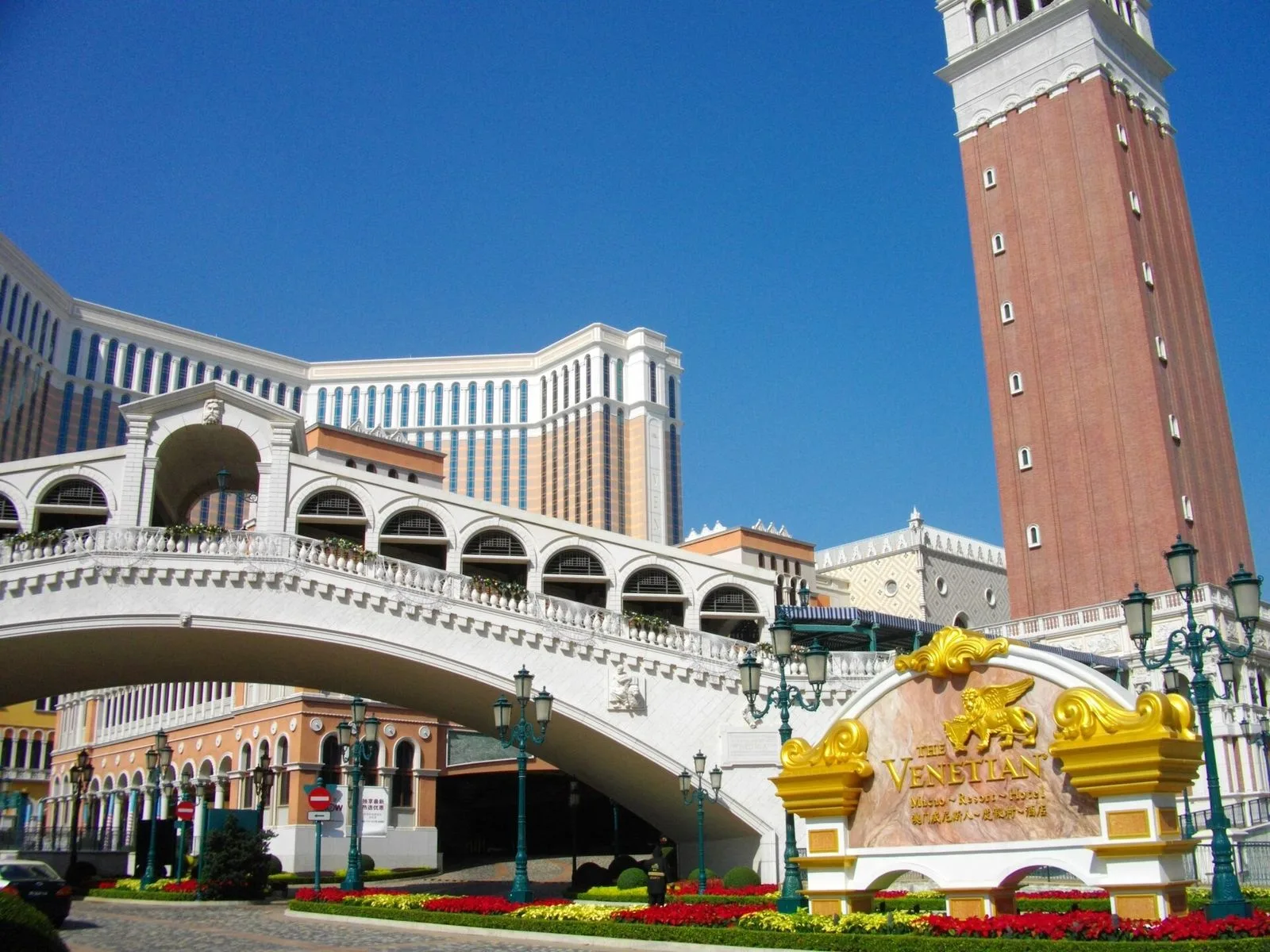 The exterior entrance of The Venetian Macao on the Cotai Strip — the entrance marked by the golden lion statues, the stone bridge over the resort lake, and the Macao-style tower of the main building, the iconic Cotai Strip view that connects The Venetian to its sister property The Parisian across the reclaimed-land strip, the Cotai Strip itself a 2km stretch of integrated resorts built on reclaimed land between Taipa and Coloane