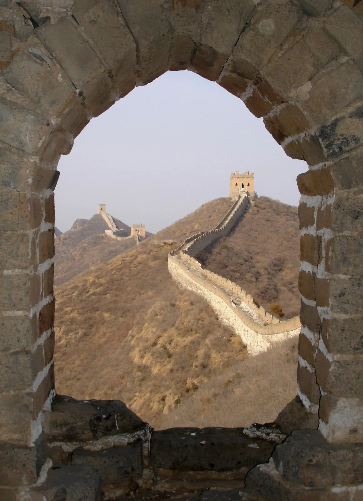 View through a watchtower window of the Great Wall snaking along a bare mountain ridge in the distance, with two more watchtowers visible on the next ridge under a hazy sky — the dramatic perspective that makes Mutianyu one of the most photographed sections of the Wall