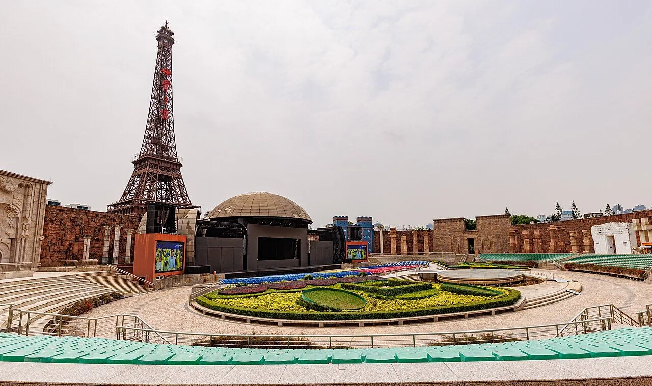 The 1:3 scale replica of the Eiffel Tower at Window of the World Shenzhen, with the outdoor amphitheater in the foreground featuring the colorful flower clock and green seating, showing the scale of the 108m replica compared to the original in Paris