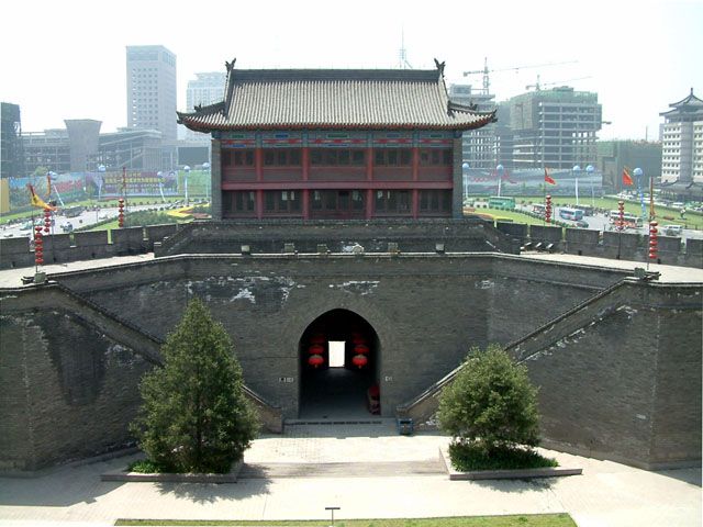 The South Gate (Yongning Gate, 永宁门) of the Xi’an city wall — the imposing Ming-dynasty gatehouse tower with grey brick walls and the red wooden upper level, the main entrance through the central archway with the red lanterns, the modern Xi’an skyline of office towers visible in the background, the most-visited of the 18 city wall gates and the historic main entrance to the ancient Tang-dynasty capital city