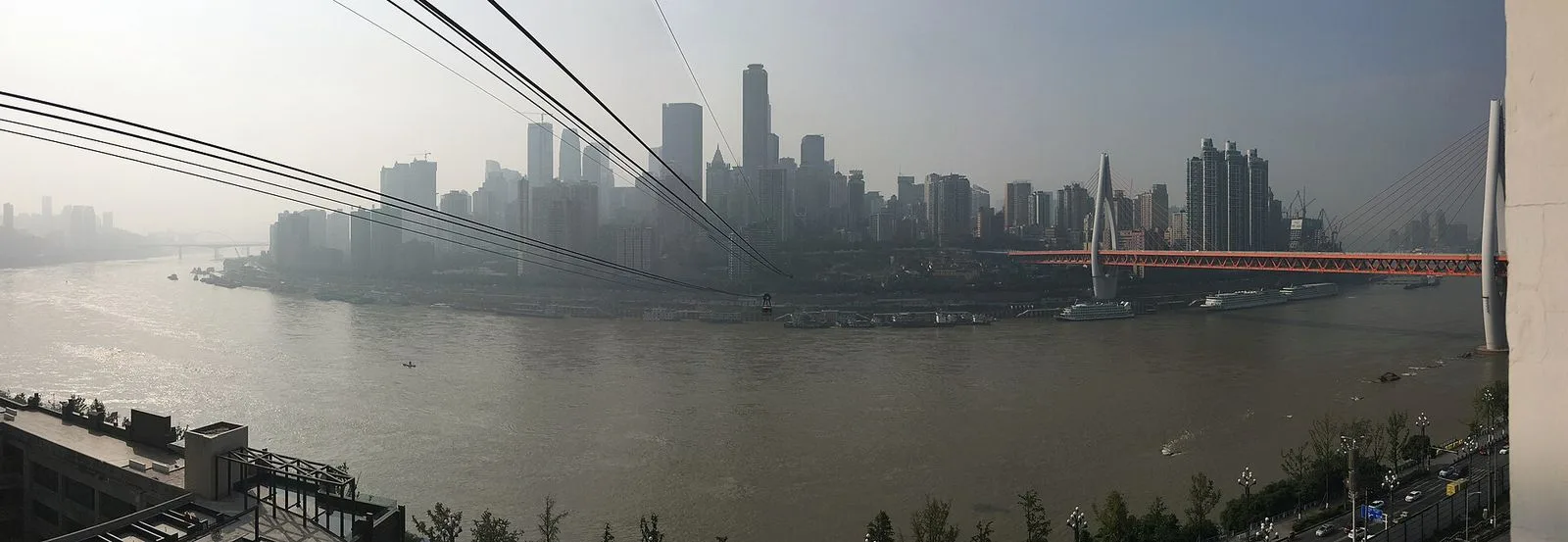 A view from inside the Chongqing Yangtze River Cableway cabin — the steel cables extending into the distance over the Yangtze River, the dramatic Chongqing skyline with its forest of skyscrapers rising above the misty river, the two modern cable car pylons painted red, the perspective the 4 million annual passengers experience on the 5-minute crossing between the Yuzhong and Nan’an districts