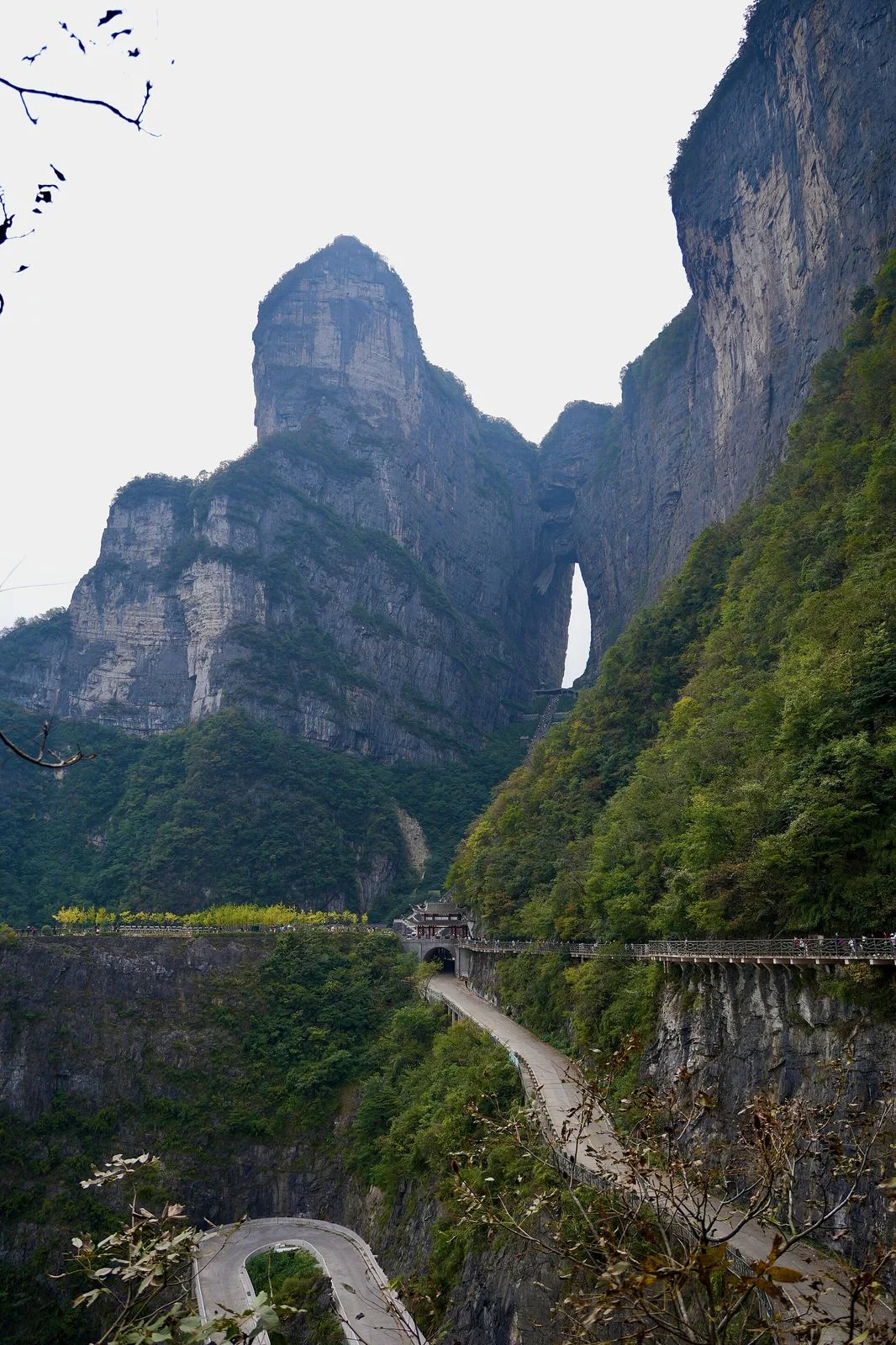A side view of Tianmen Mountain showing the famous Tianmen Cave (Heaven’s Gate) natural arch and the dramatic mountain peaks of Zhangjiajie National Forest Park — the steep cliffs of the mountain and the small hole of the cave visible at the top center, the 99-bend mountain road (99道弯) winding up the mountain at the bottom right, the overview shot that shows the 1,518m mountain rising from the Hunan countryside, the location of the world’s longest passenger cableway
