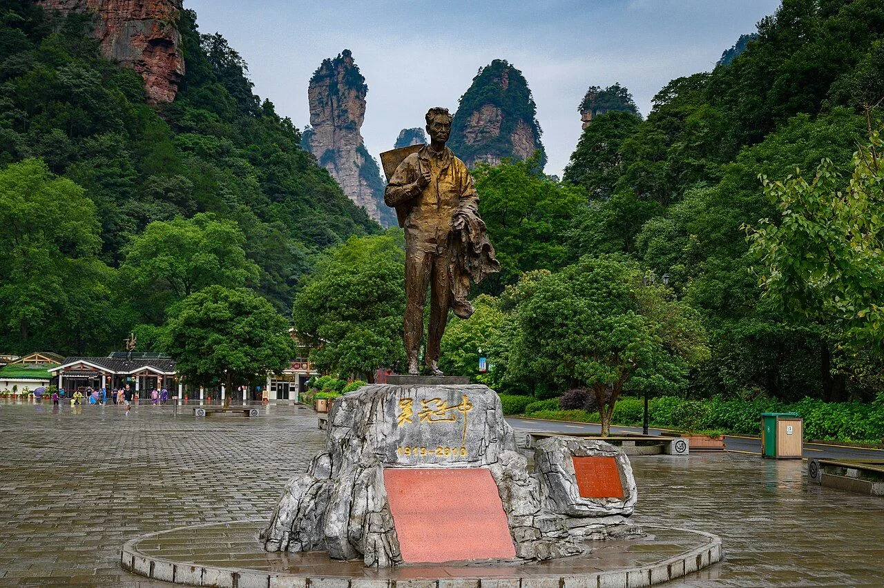 The bronze statue of painter Wu Qianqiu (1917-1999) at the entrance plaza of Zhangjiajie National Park, a giant standing figure holding his paint case, with the dramatic sandstone pillar peaks rising behind him in the mist and a few visitors walking in the rainy plaza