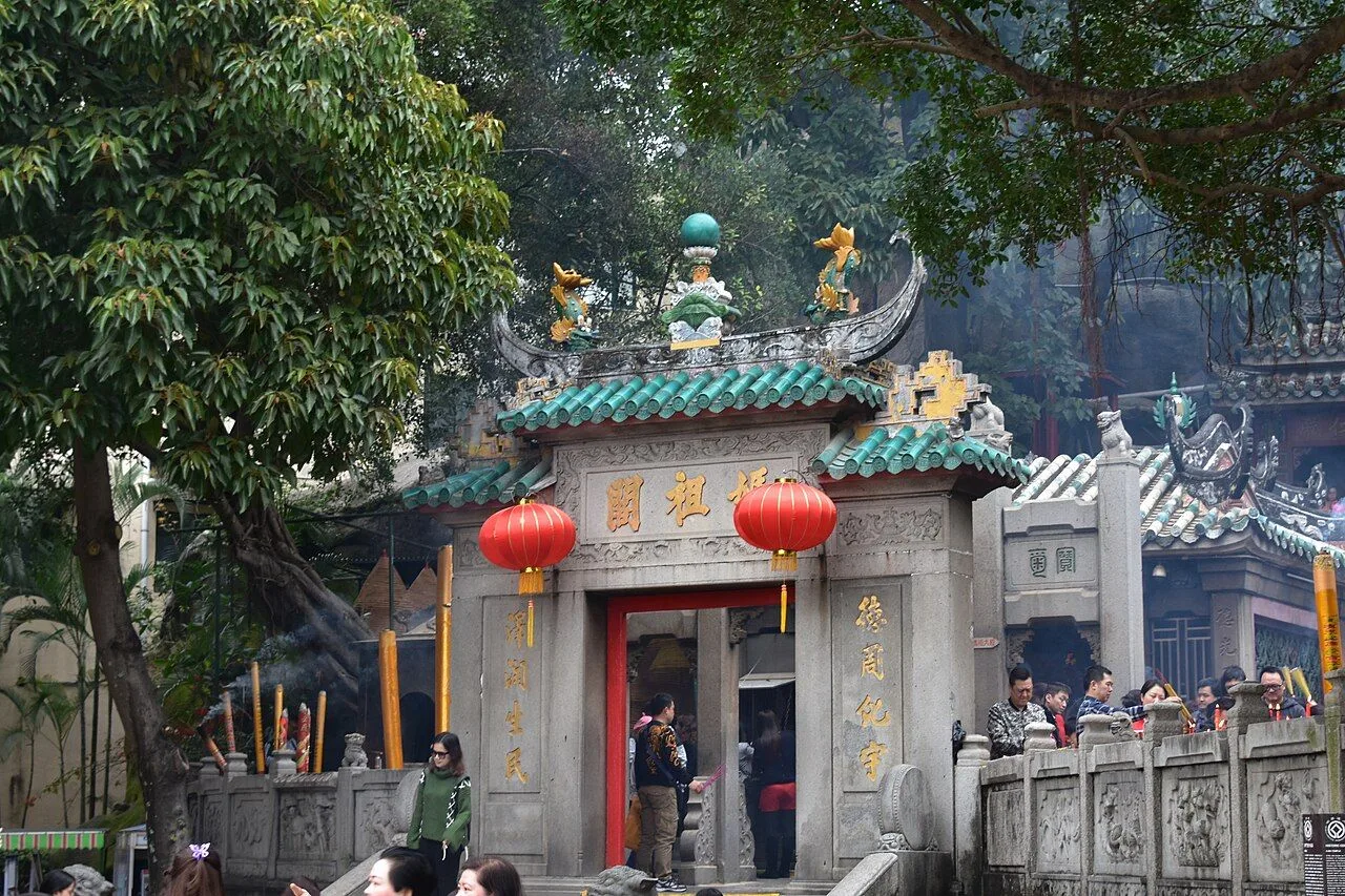 The prayer hall gate of the A-Ma Temple in Macau, with the characters 天后聖母 (Holy Mother of Heaven) on the lintel, red lanterns, and local worshippers arriving for morning prayers