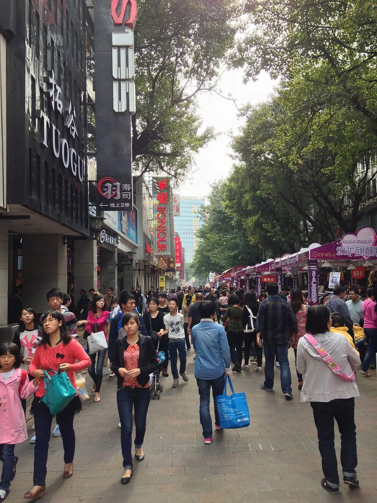 Daytime wide view of Beijing Road pedestrian street showing the 1930s arcade buildings on both sides, the modern storefronts, and the canopy of mature trees providing shade