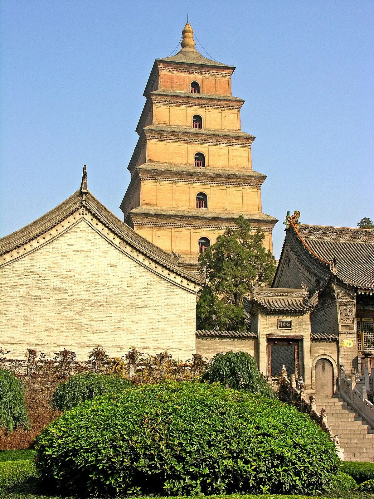 The Big Wild Goose Pagoda in Xi'an seen from the temple gardens, with a traditional grey-brick temple building in the foreground and manicured shrubs framing the base of the 1,370-year-old Tang-brick tower