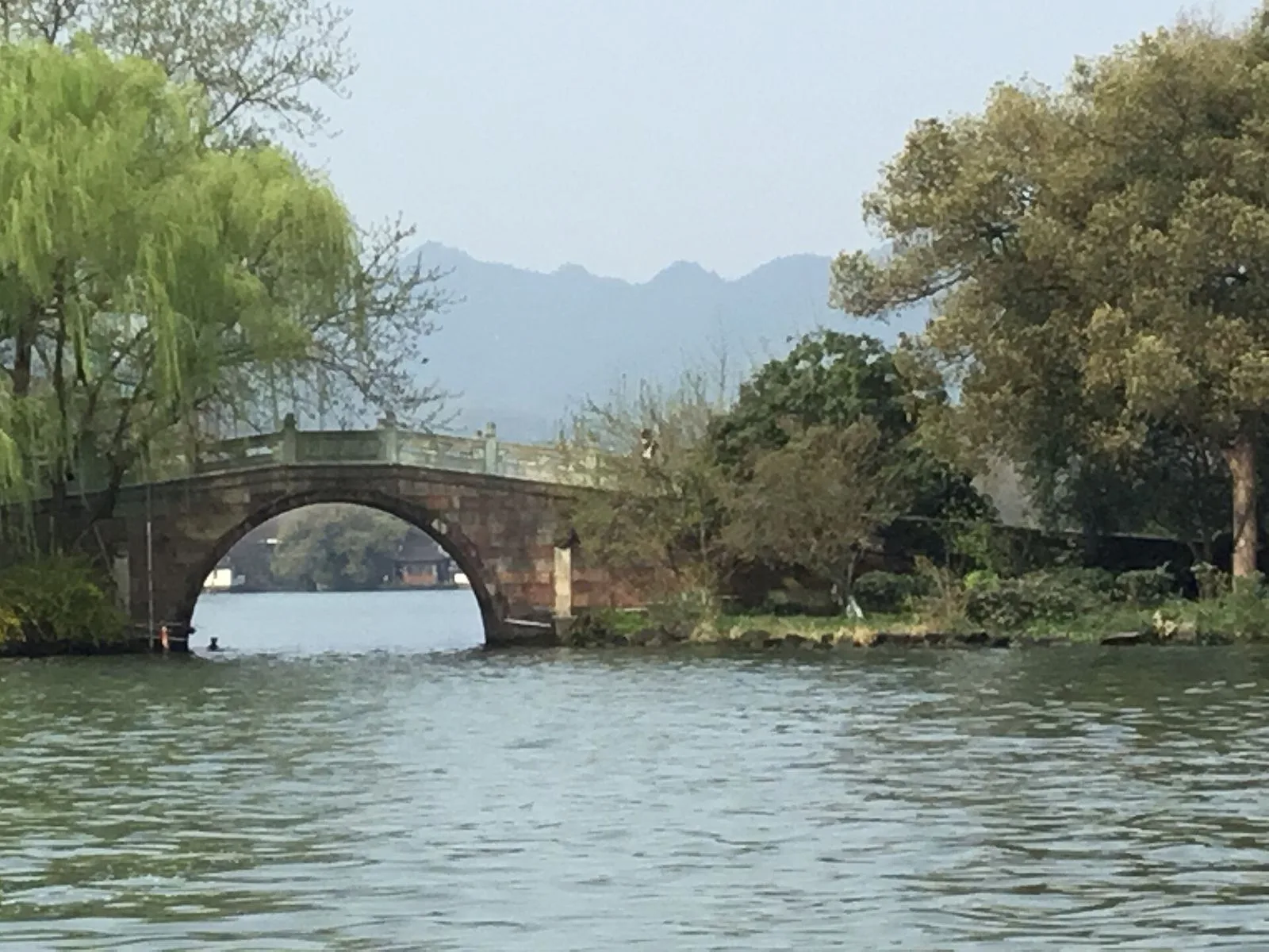 A traditional arched stone bridge over the calm waters of West Lake, framed by weeping willow trees on both sides, with a faint mountain silhouette in the hazy distance — the timeless Jiangnan landscape that has inspired Chinese poetry and painting for over 1,000 years, the same scene referenced in the famous "Ten Scenes of West Lake" poems