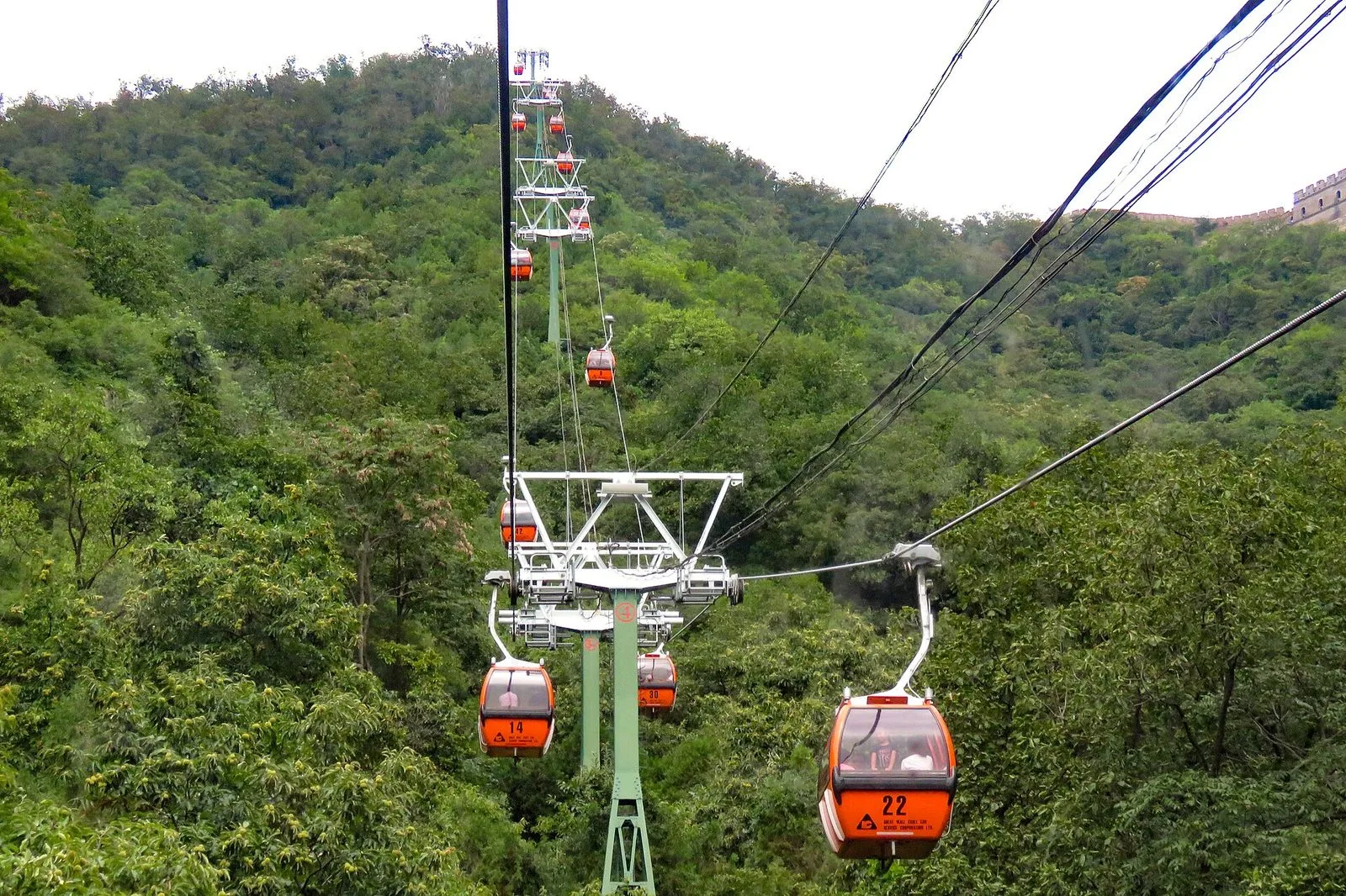 Orange cable car gondolas ascending the forested mountain above Mutianyu, with cables stretching from the valley floor up to the ridge where the Wall sits — the most popular way to get up to the wall, taking 20 minutes round trip and avoiding the 1,000+ step climb in summer heat