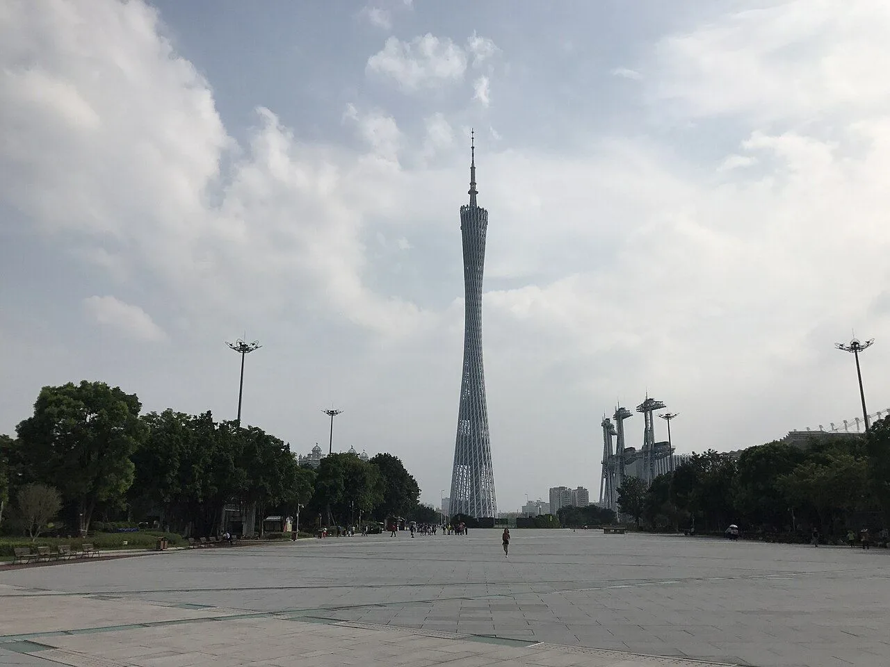 Ground-level view of Canton Tower in Guangzhou, looking up the broad plaza with the tower rising into the cloudy sky, and visitors walking toward the base — the pedestrian approach to the third-tallest TV tower in the world