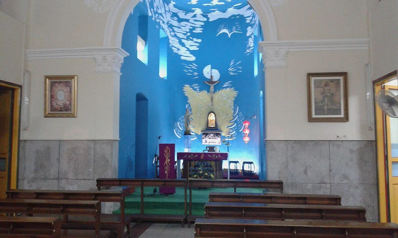 The interior of the St. Francis Xavier Chapel in Coloane Village, Macau, with the colorful Magnificat mosaic side altar (the Canticle of Mary in English, Portuguese, and Chinese), wooden pews, and a wall-mounted electric fan — the chapel in continuous use since the 17th century