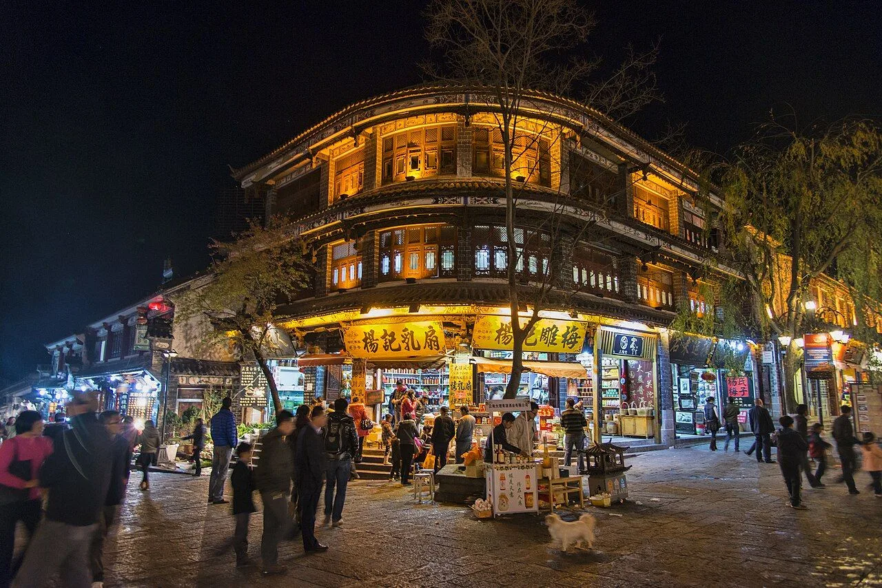 A corner shop lit up at night in Dali Old Town, with the three-story wooden traditional Bai architecture, locals shopping, and a small dog in the foreground — the lively old town at night