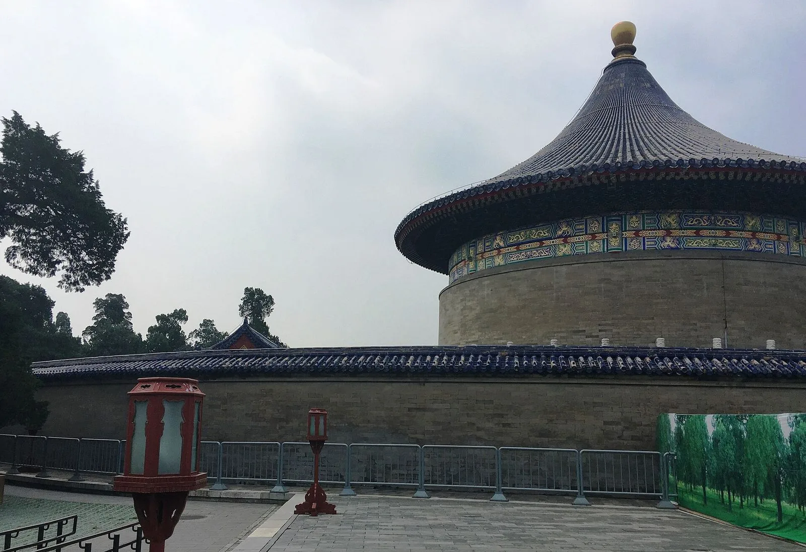 The Echo Wall at the Temple of Heaven — the curved exterior of the Imperial Vault of Heaven's circular wall with its blue-glazed tile roof and ornate painted beams, a traditional red lantern standing in the foreground on the stone-paved walkway, and the Imperial Vault of Heaven visible behind. The 65-meter circular wall is famous for its acoustic property that carries a whisper from one end to the other