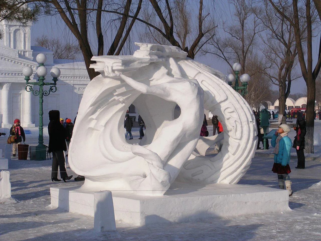A circular ring-form snow sculpture at the Harbin festival with visitors in winter clothing and pine trees in the background — the scale of the snow sculptures (multiple meters tall) and the -25°C weather give a sense of the festival