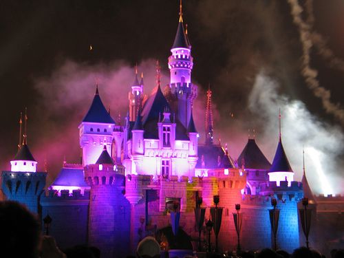 The Sleeping Beauty Castle at night lit up in blue and purple with fireworks smoke — the iconic Disney night scene, the castle transforms colors during the evening fireworks and light show