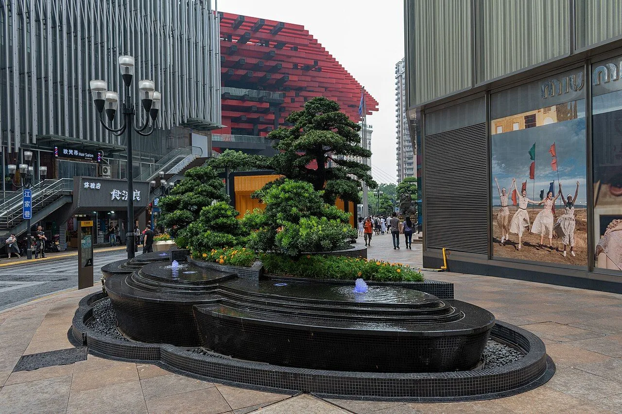 The Jiefangbei Pedestrian Street with a black stone fountain and bonsai tree, a Miu Miu billboard, the iconic Jiefangbei red metal sculpture, and locals walking through — daytime shopping scene at one of the 5 malls in the area