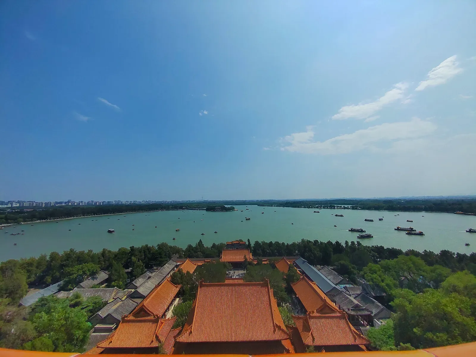 Kunming Lake at the Summer Palace, viewed from the top of Longevity Hill — the vast lake stretches into the distance with tour boats, traditional pavilions on the hillside including the Tower of Buddhist Incense, and the modern Beijing skyline barely visible through the haze on the horizon