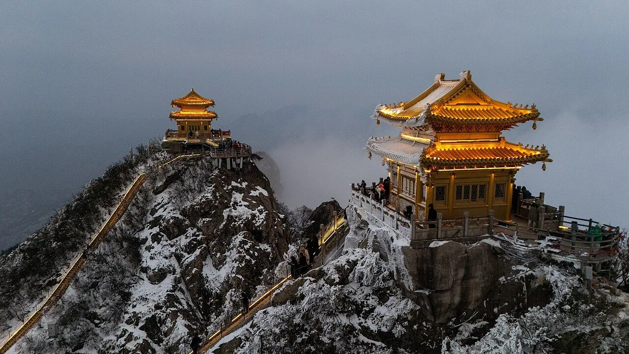 Two illuminated golden-roofed Taoist temples on the snowy peaks of Laojun Mountain at dusk — a glowing walkway between them, clouds of fog rolling in below, and a visitor standing at the railings; the most photogenic winter scene in central China, at 2,217m elevation on the Funiu Mountains