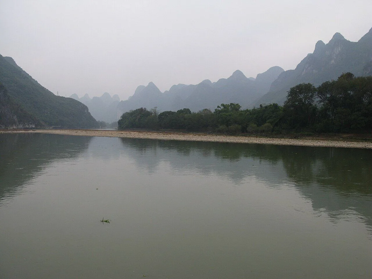 The Li River at its most iconic moment — a perfect mirror reflection of the karst peaks and overcast sky on the still water, with a curved gravel bar in the foreground; the landscape depicted on the back of the 20-yuan banknote and the inspiration for countless Chinese landscape paintings